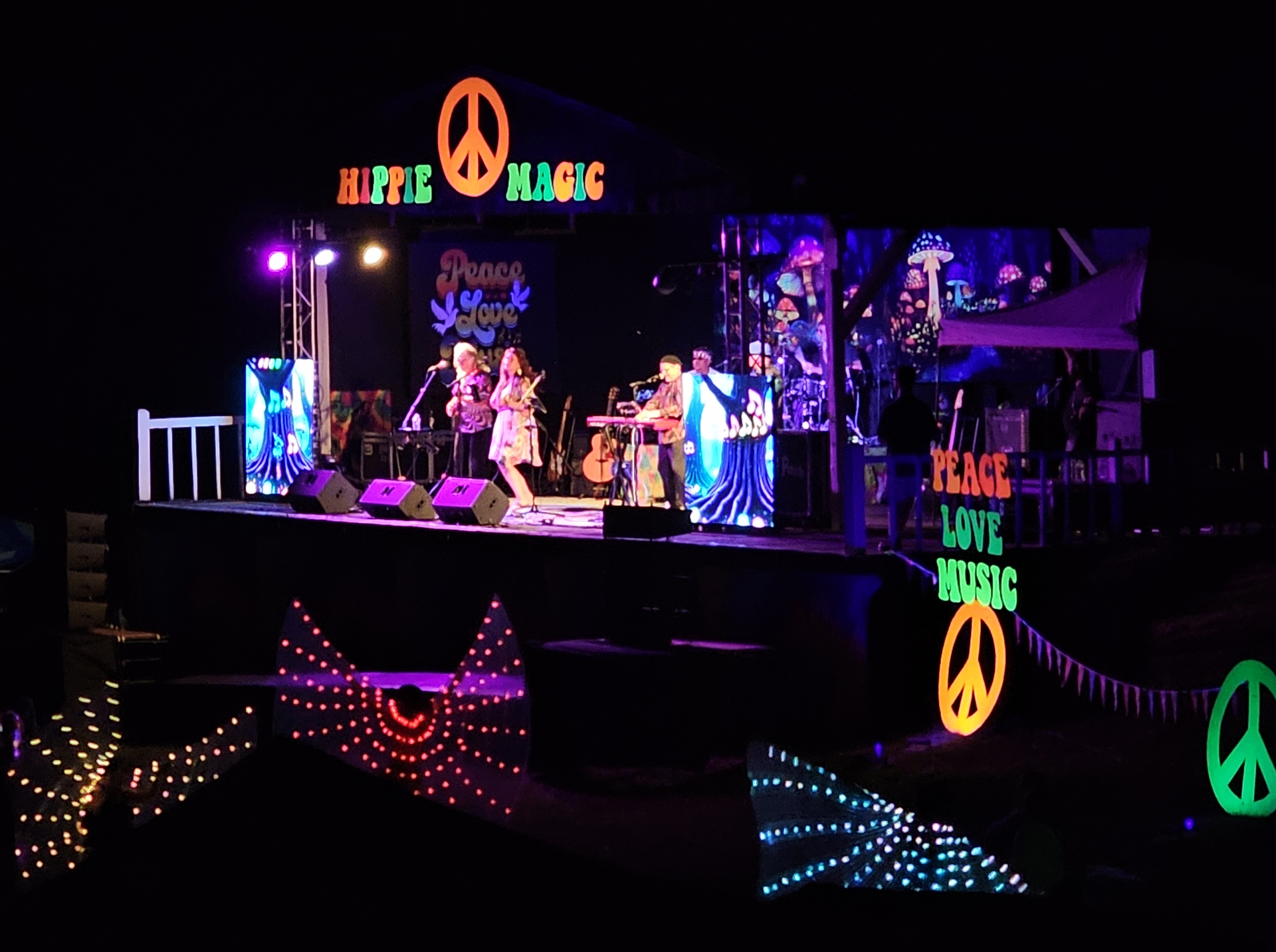 HIPPIE MAGIC main stage lit up at night with neon peace signs and a live band